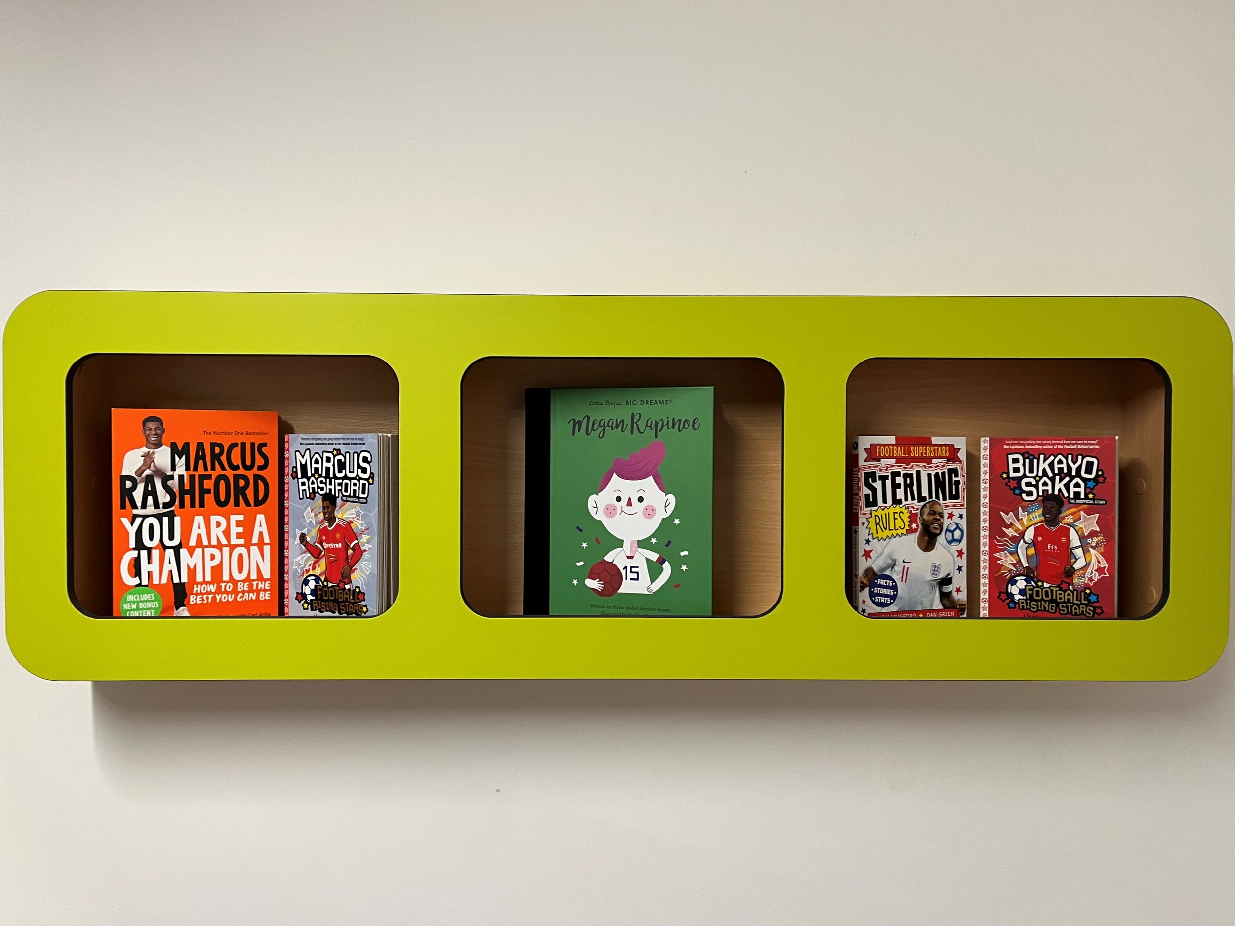 A green wall-mounted shelf with three compartments displaying books. The left compartment contains two Marcus Rashford books. The middle compartment has a book about Megan Rapinoe. The right compartment features books on Sterling and Bukayo Saka.