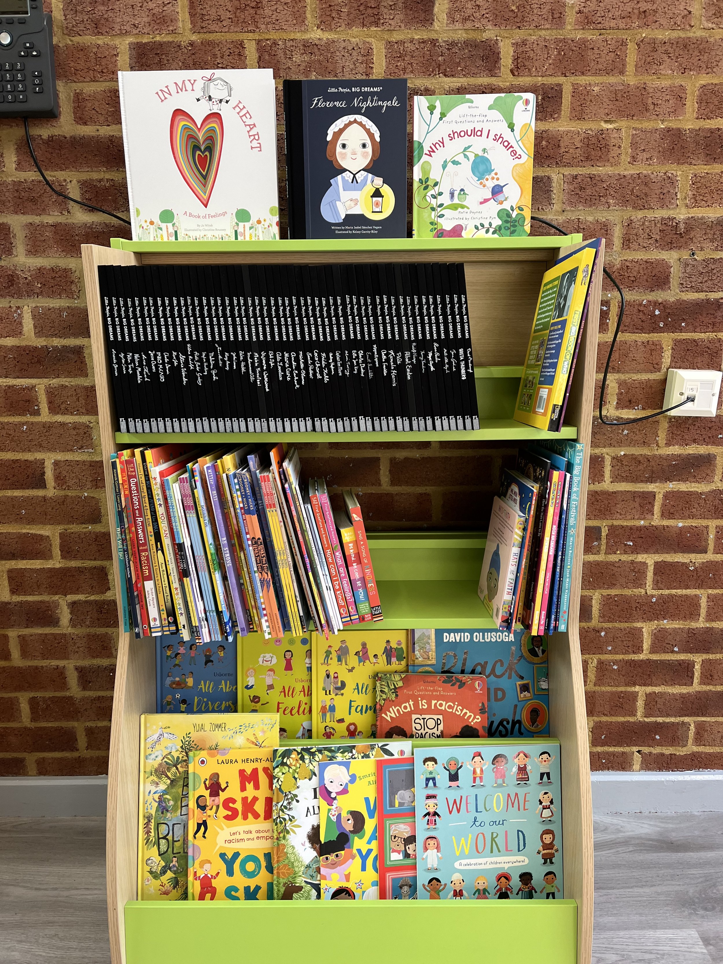 Children's book display with shelves against a brick wall, showcasing various titles including "In My Heart," "Florence Nightingale," and "Welcome to Our World."